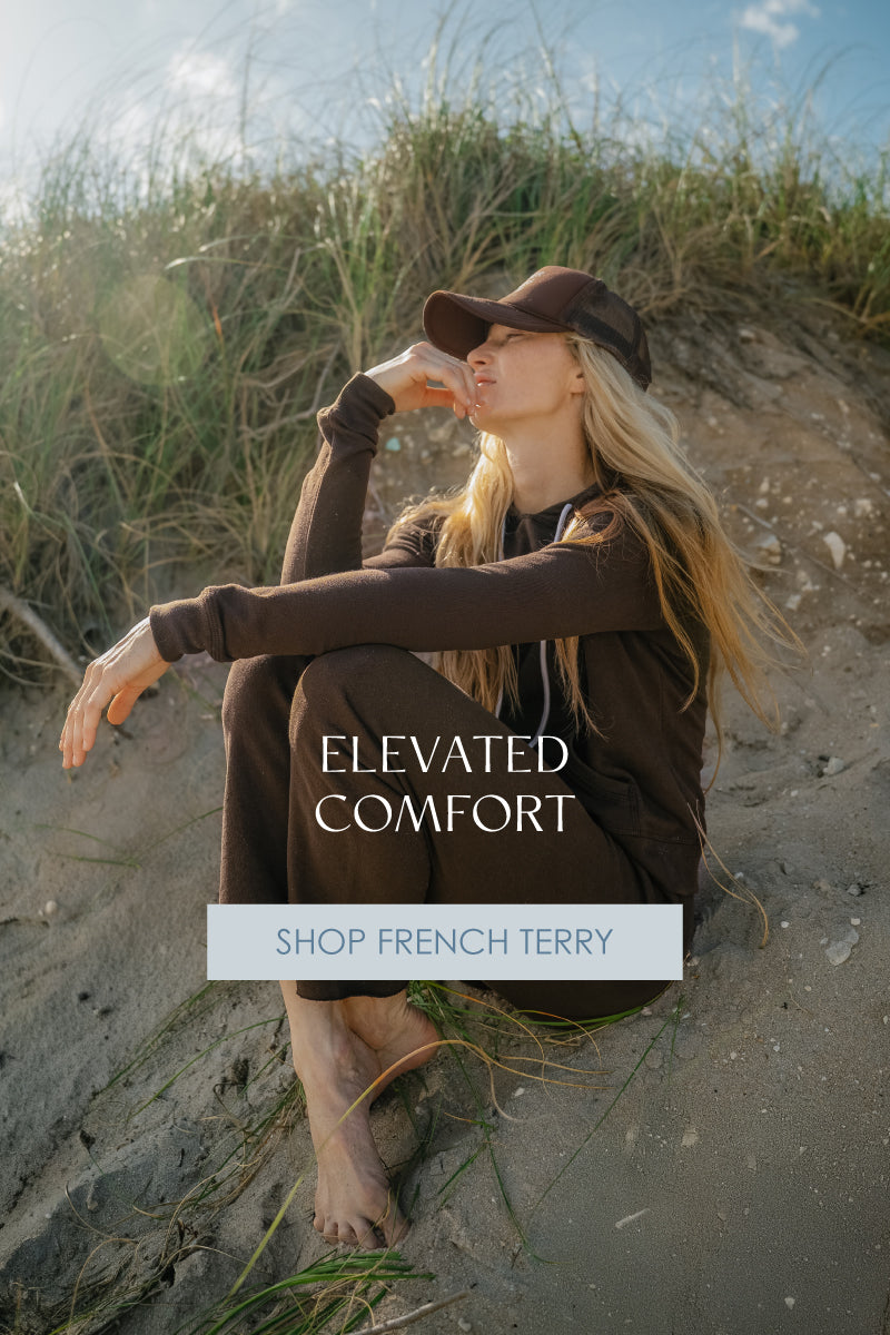 Model wearing French Terry hoodie and pants in Espresso on beach dune; headline reads ‘Elevated Comfort.’