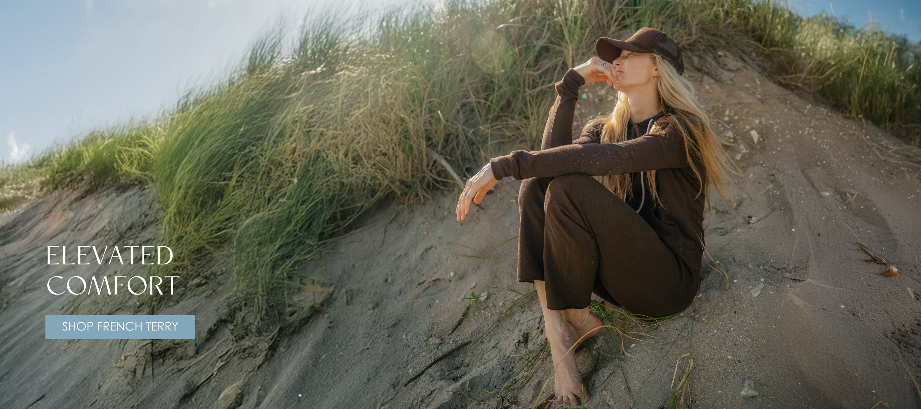 Model wearing French Terry hoodie and pants in Espresso on beach dune; headline reads ‘Elevated Comfort.’