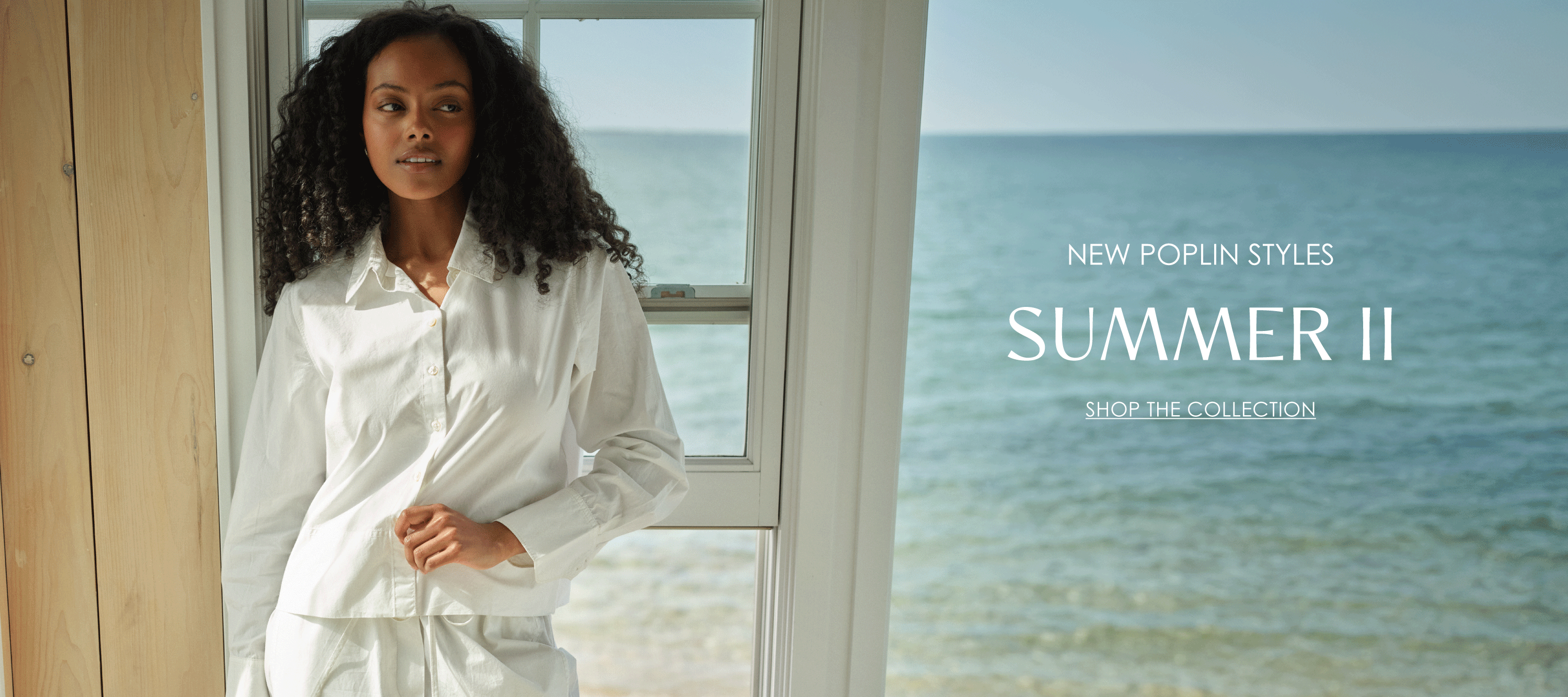 New Poplin Styles. Summer II. Shop the Collection. Model wearing the Laurel Button Down in White, styled with matching poplin pants, standing by a window overlooking the ocean.